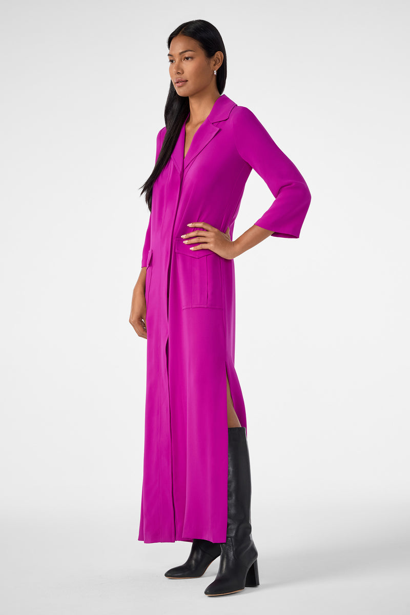 A woman models the Annecy Dress—a magenta, long-sleeve, collared button-down with front pockets and side slits—paired with knee-high black boots. She poses with one hand on her hip against a plain white background.