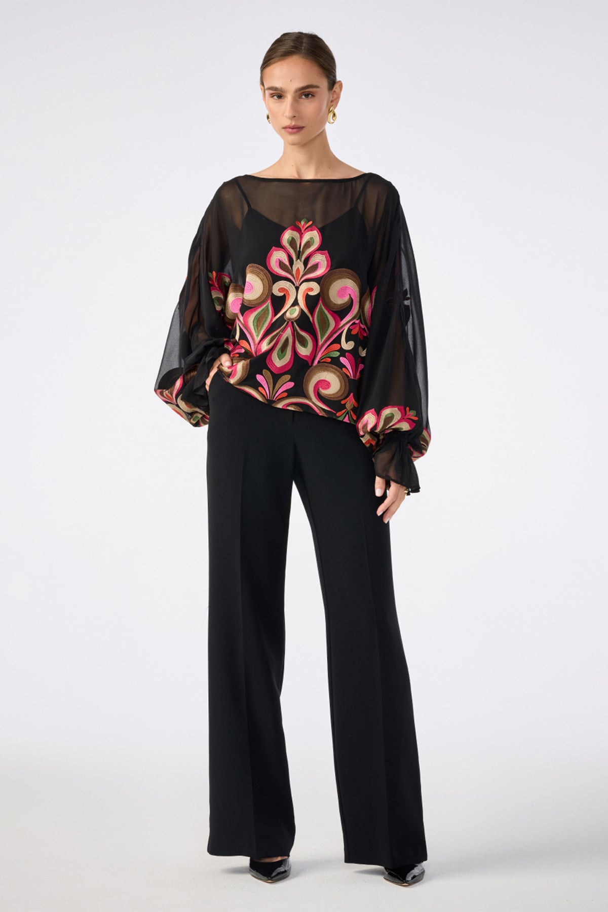 A woman wears the Soho Embroidered Top by Sanctuary—a sheer black blouse with voluminous sleeves and a bold, colorful floral front—paired with black wide-leg pants, hoop earrings, and black pointed shoes against a plain white background.