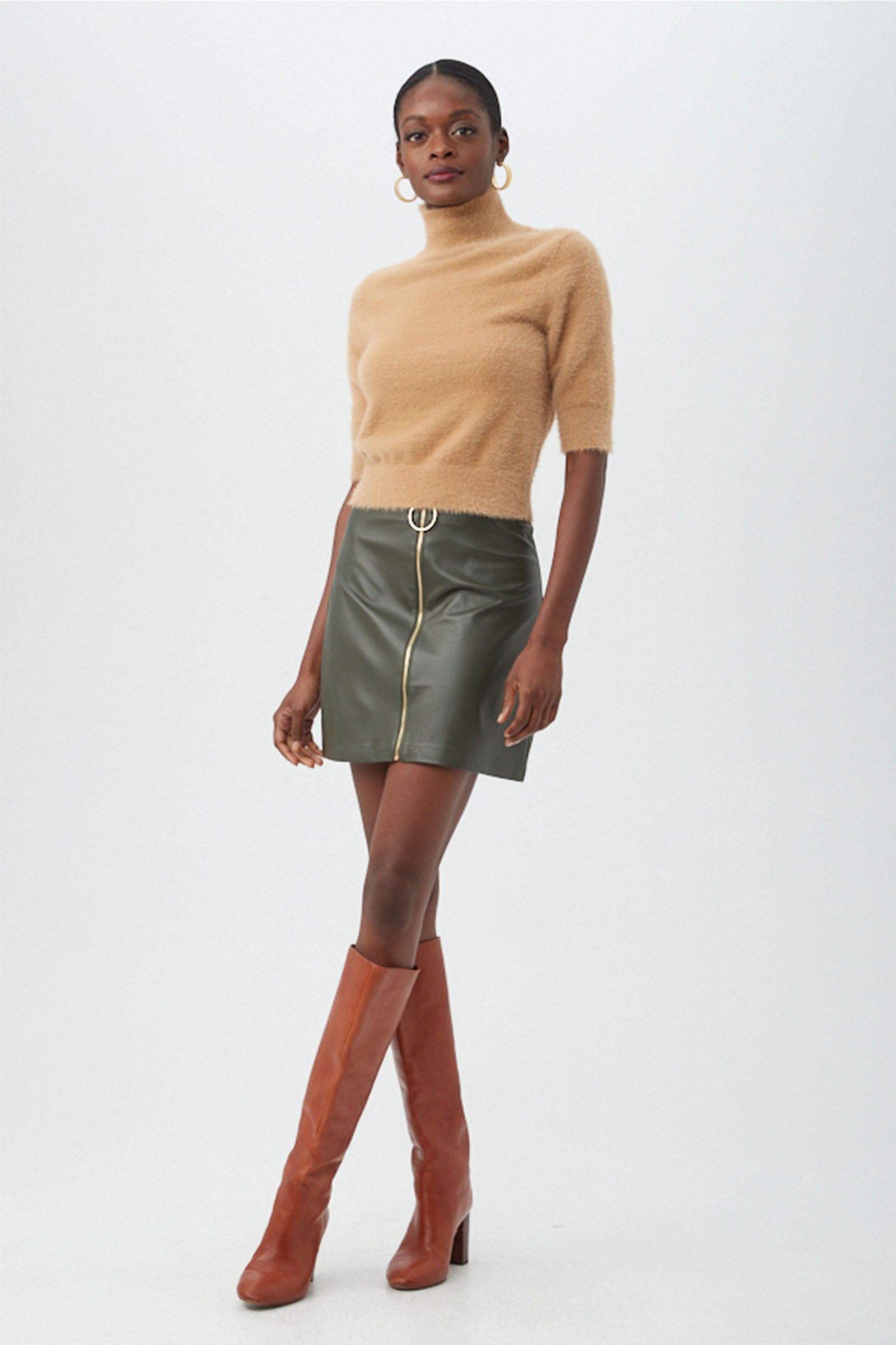A woman poses in the Mali Fuzzy Sweater, a beige short-sleeve turtleneck, paired with an olive green leather mini skirt featuring a front zipper, tall brown boots, and large hoop earrings.