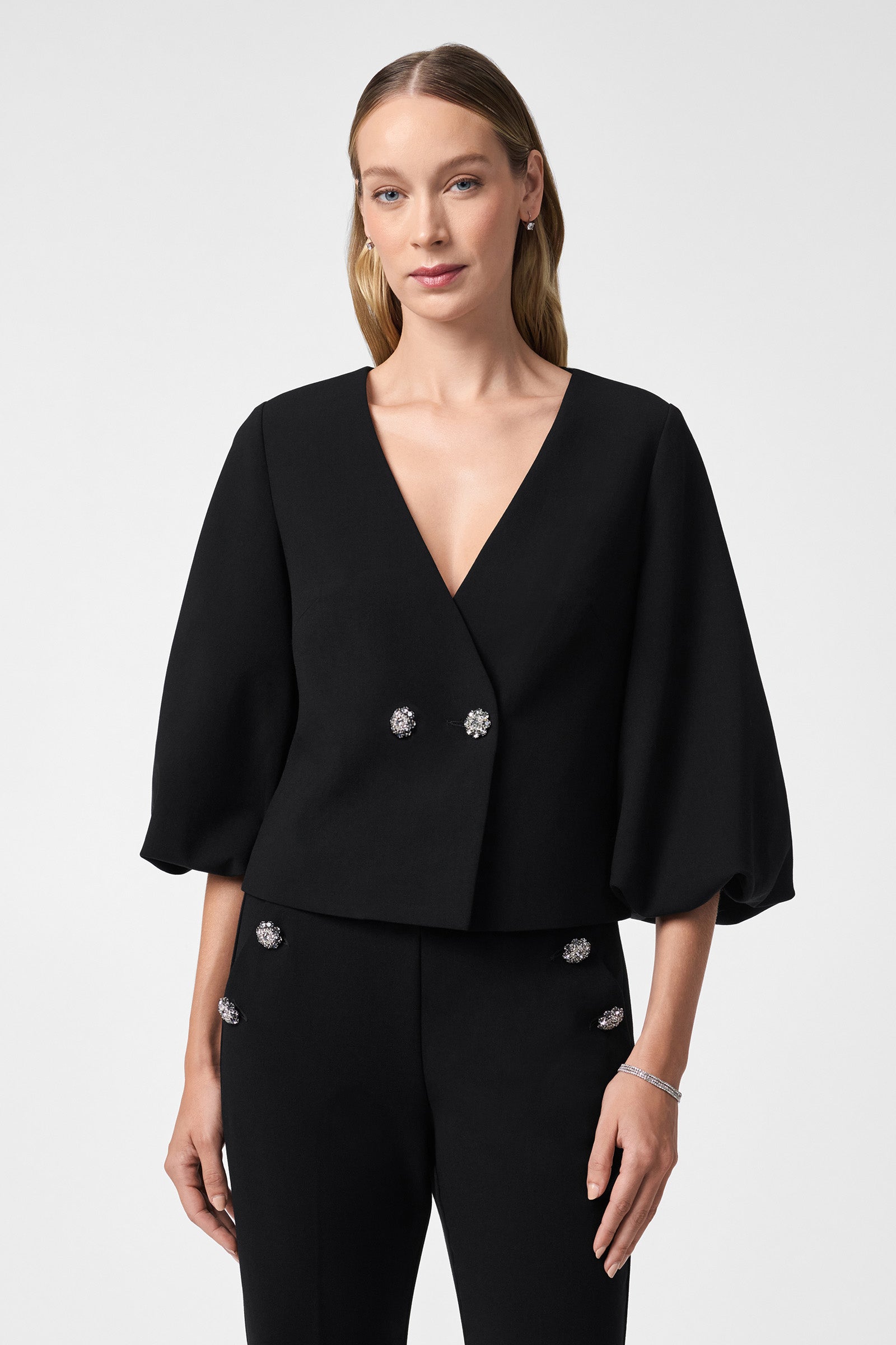 A woman with long blonde hair smiles slightly, wearing the Basal Cropped Jacket—a black V-neck top with wide three-quarter sleeves and jeweled buttons—paired with matching black pants, posed against a plain light background.
