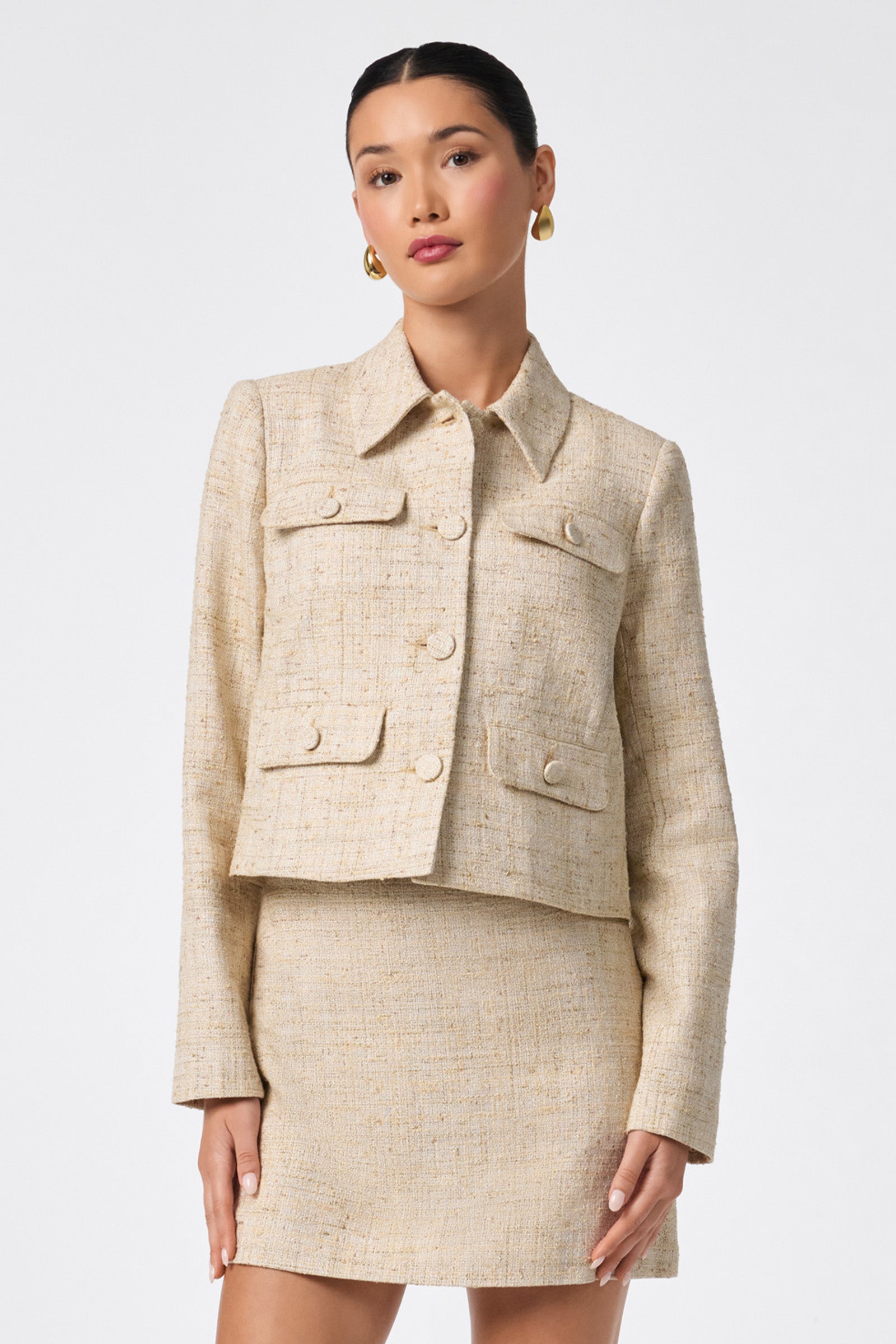 A woman stands against a white background wearing the Bayonne Jacket, a light beige tweed piece with front pockets and large buttons, paired with a matching skirt, her hair slicked back and accessorized with gold hoop earrings.