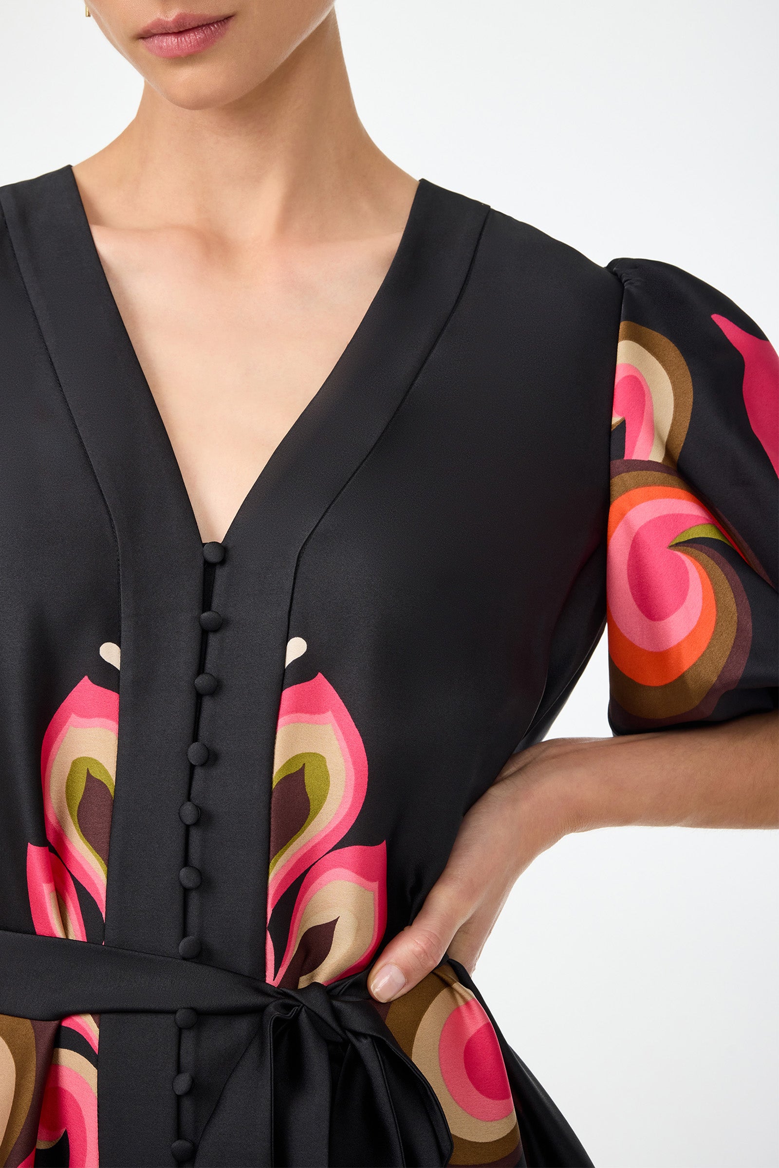 A woman wears the Certonia Puff Sleeve Dress - Sanctuary, featuring a black base, deep V-neck, fabric-covered buttons, waist tie, and colorful abstract pink, orange, and beige patterns on the sleeves and bodice. Her right hand rests on her hip.