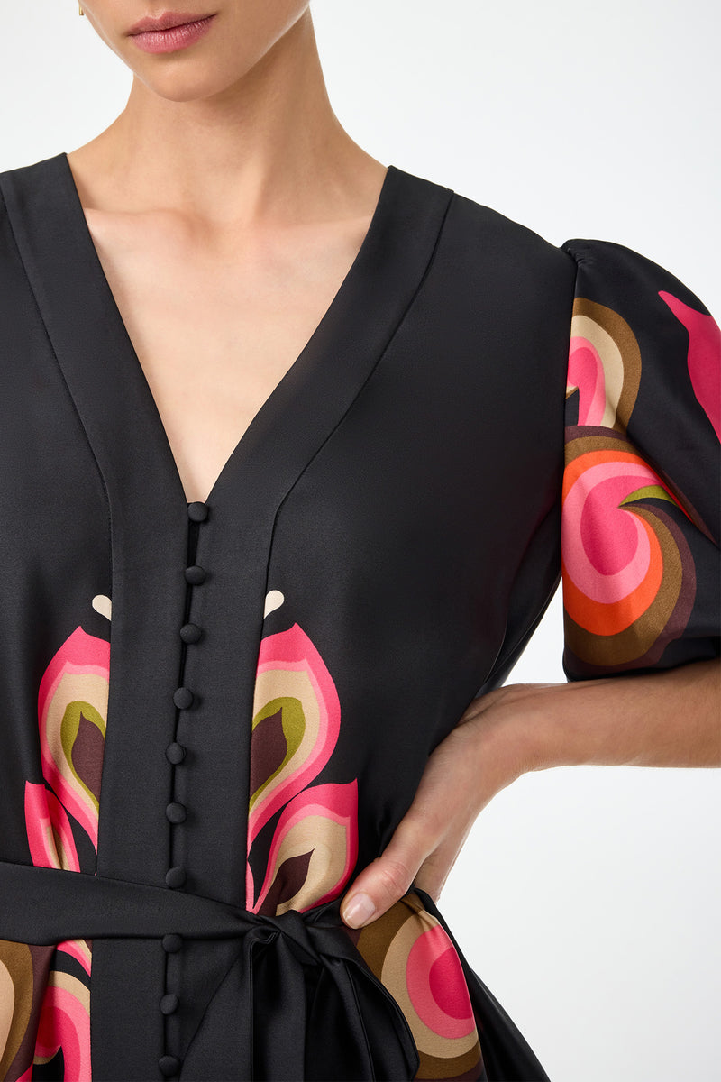 A woman wears the Certonia Puff Sleeve Dress - Sanctuary, featuring a black base, deep V-neck, fabric-covered buttons, waist tie, and colorful abstract pink, orange, and beige patterns on the sleeves and bodice. Her right hand rests on her hip.