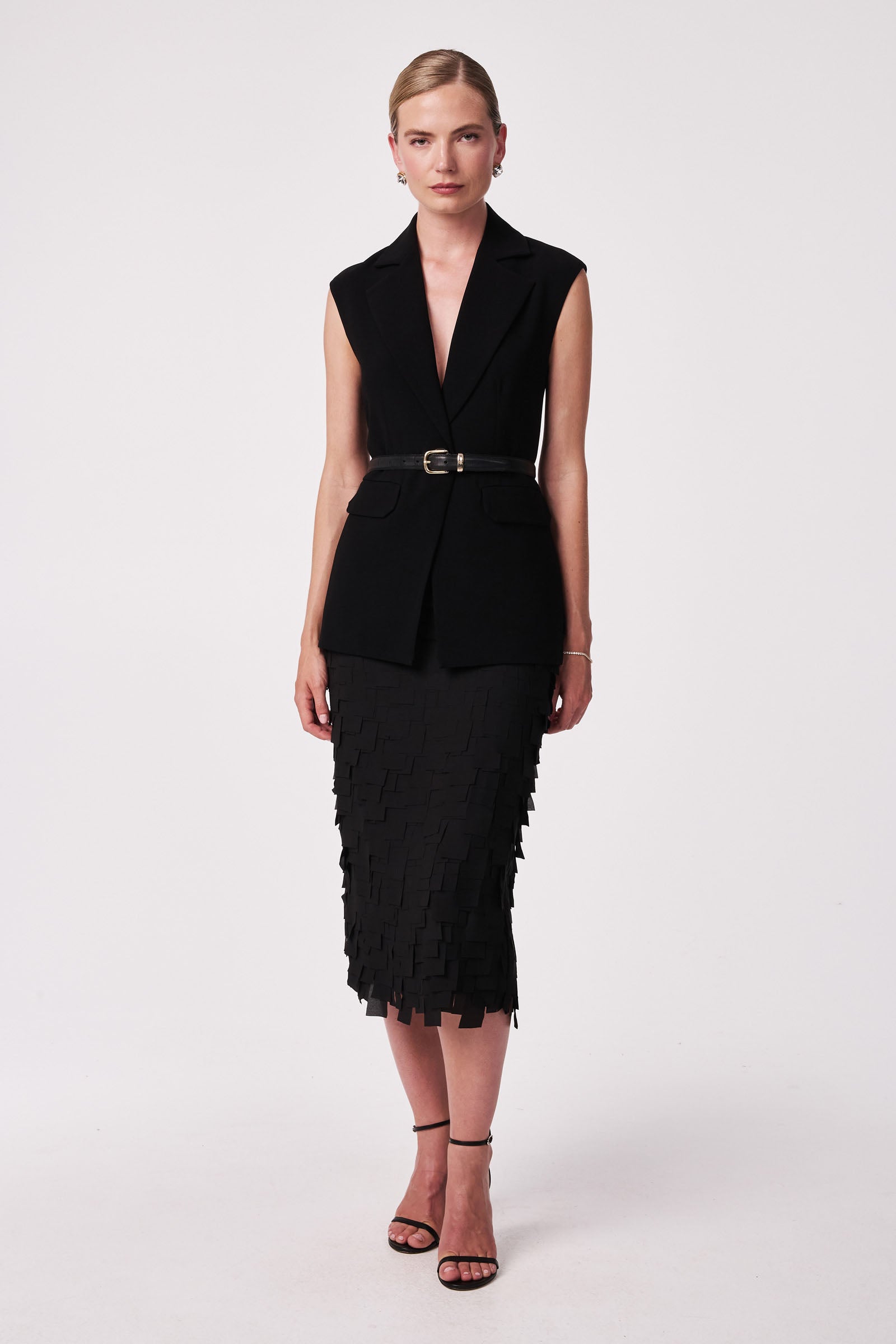 A woman faces forward in the Dita Midi Pencil Skirt - Flutter Chiffon, a black sleeveless belted blazer, and black heeled sandals, standing against a plain white background.