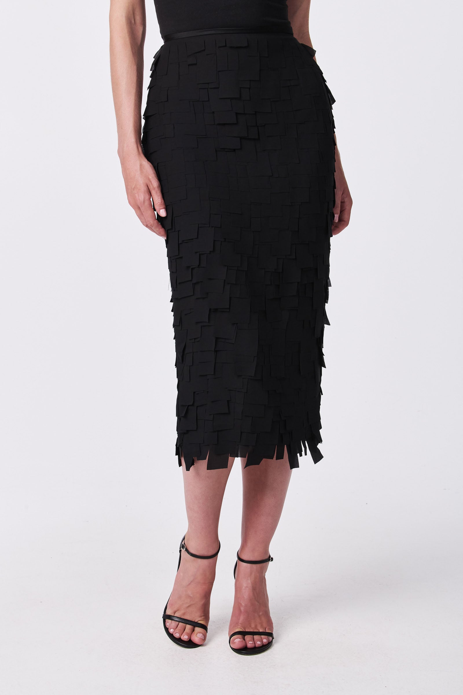A person wears the Dita Midi Pencil Skirt - Flutter Chiffon, a black, knee-length skirt with a layered, textured square pattern, paired with black high-heeled sandals against a plain white background.