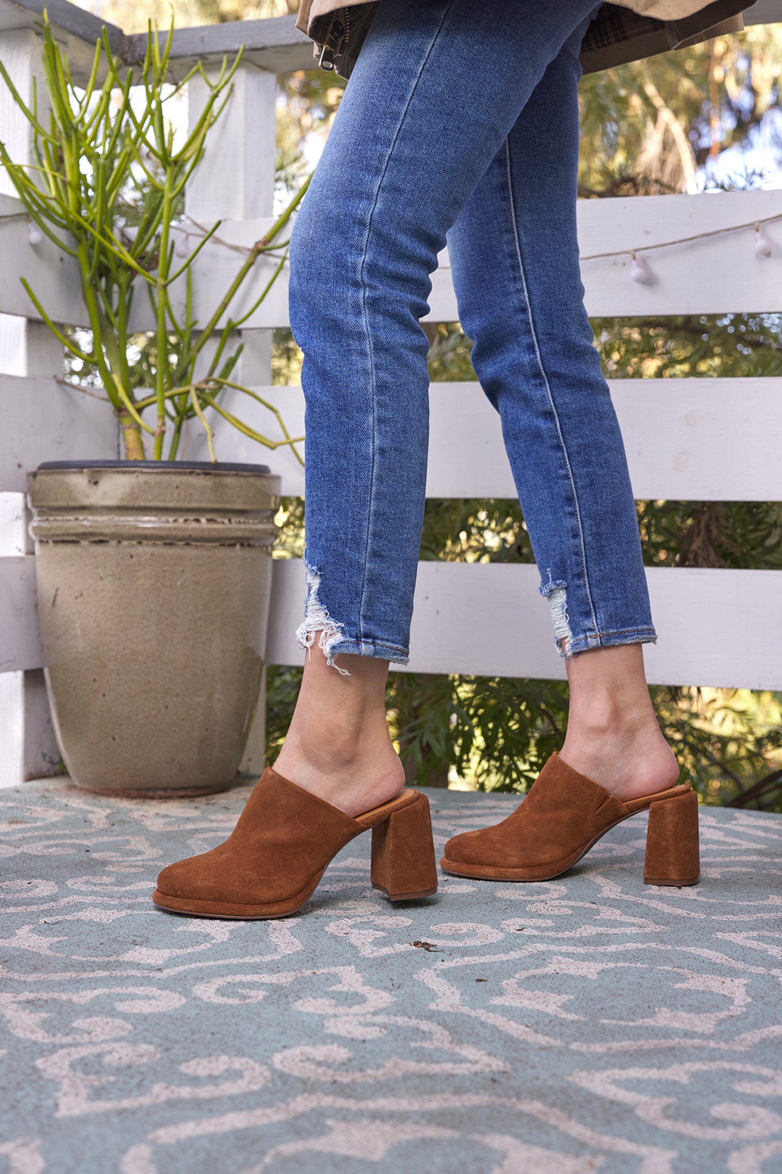 A person in blue jeans and the Honor Mule - Brown Suede stands on a patterned outdoor rug next to a large potted plant by a white wooden fence.
