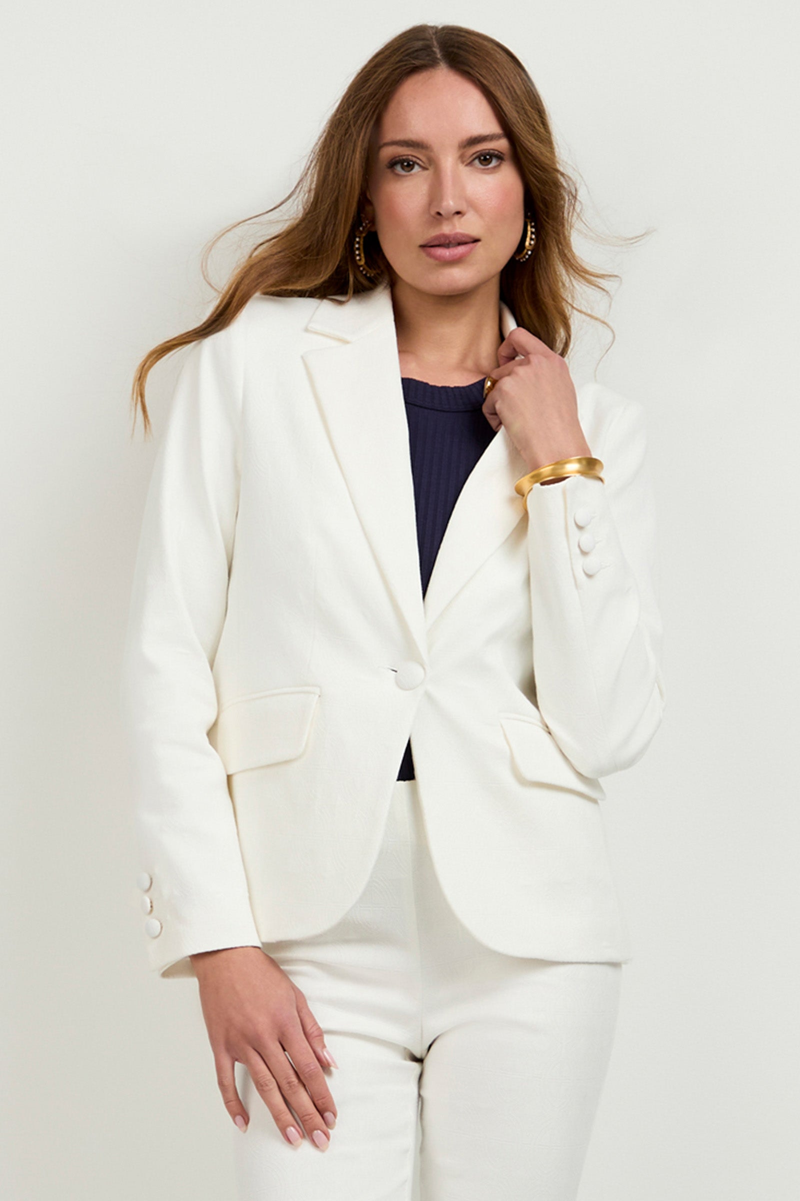 A woman with long brown hair models the Incanta Fitted Blazer and matching pants over a dark top, accessorized with gold hoop earrings and a gold bangle, posing before a plain light background.