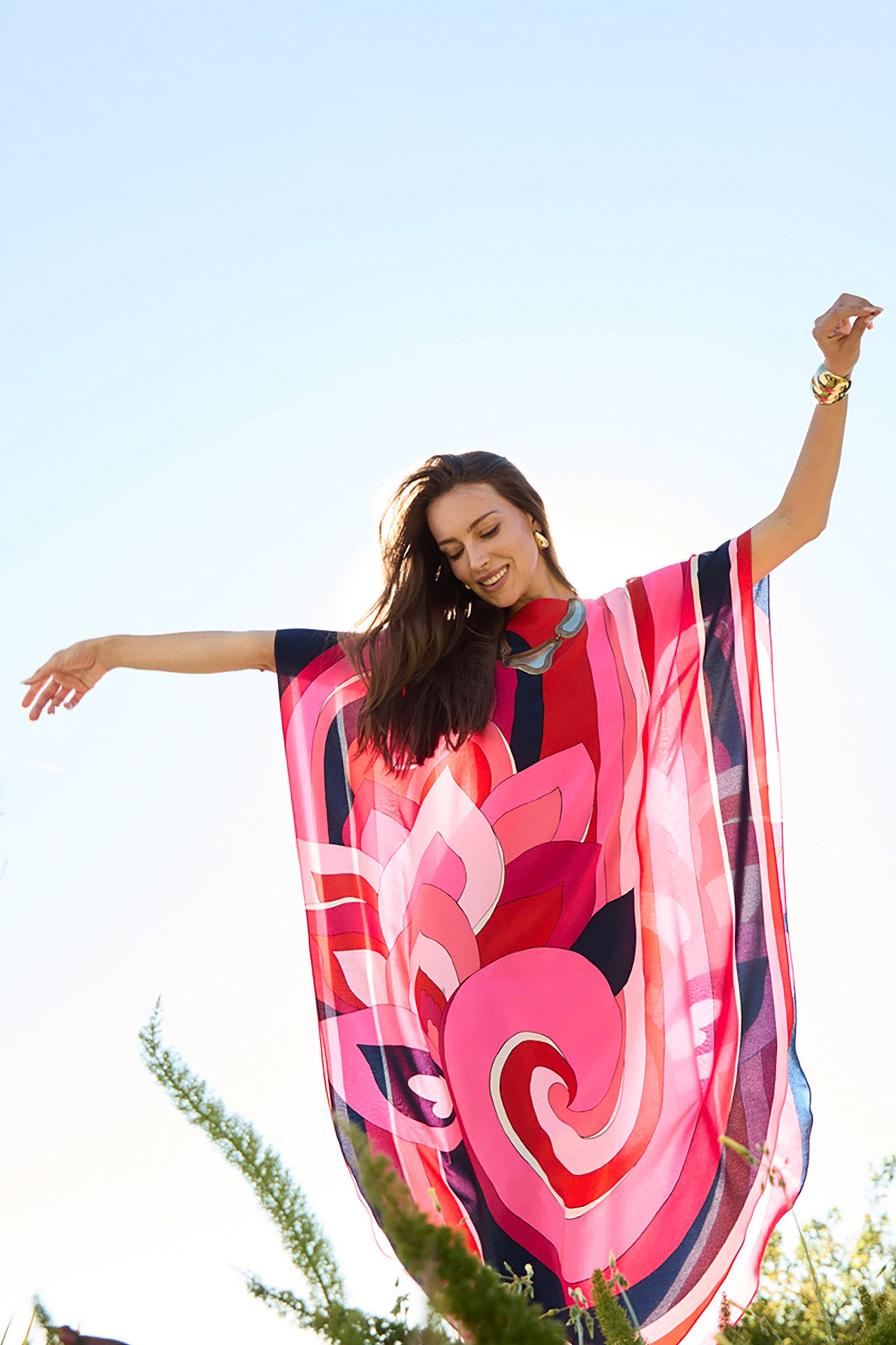 A smiling woman with long brown hair poses outdoors under a clear blue sky, arms spread wide, wearing the vibrant Jalani Maxi Caftan Dress in flowing pink and red patterns.