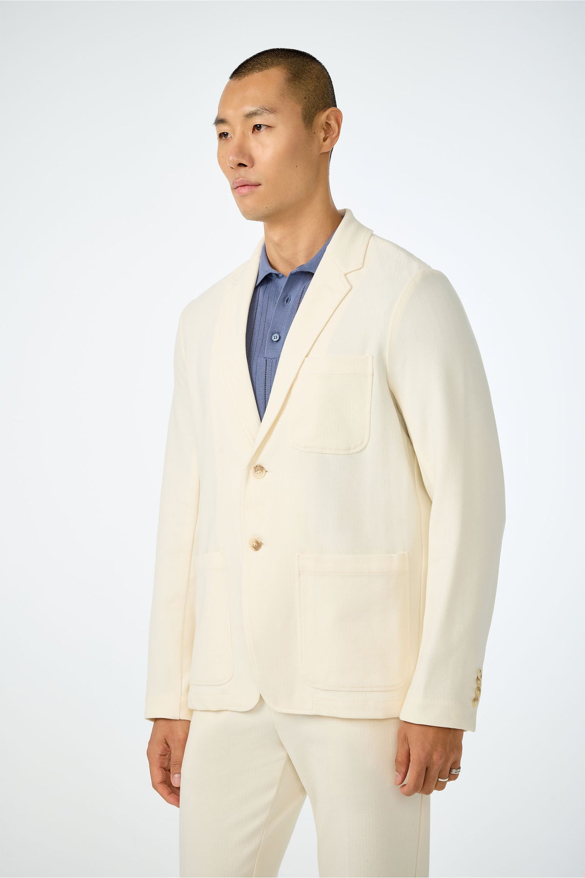 A man with short hair wears the cream-colored Muir Knit Blazer with patch pockets over a blue collared shirt, standing against a plain white background.