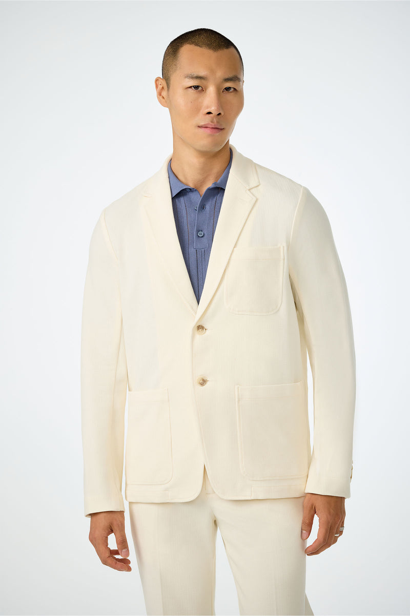 A man with short hair wears the Muir Knit Blazer in ecru over a blue collared shirt, standing against a plain light background.