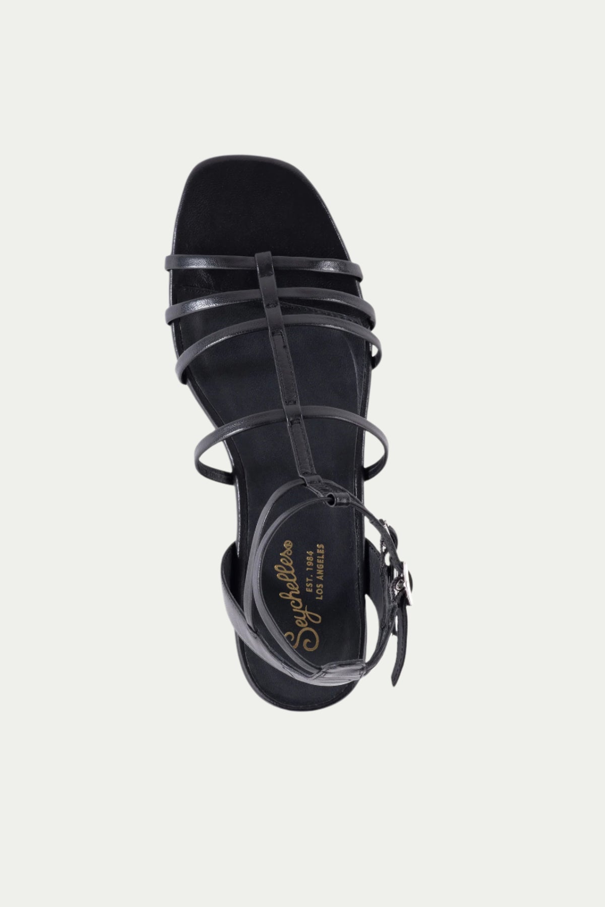 Top view of the Luxurious Wedge Sandal in black leather featuring strappy design, low wedge heel, ankle strap with buckle closure, and gold Seychelles Los Angeles branding on the insole. Set against a light gray background.