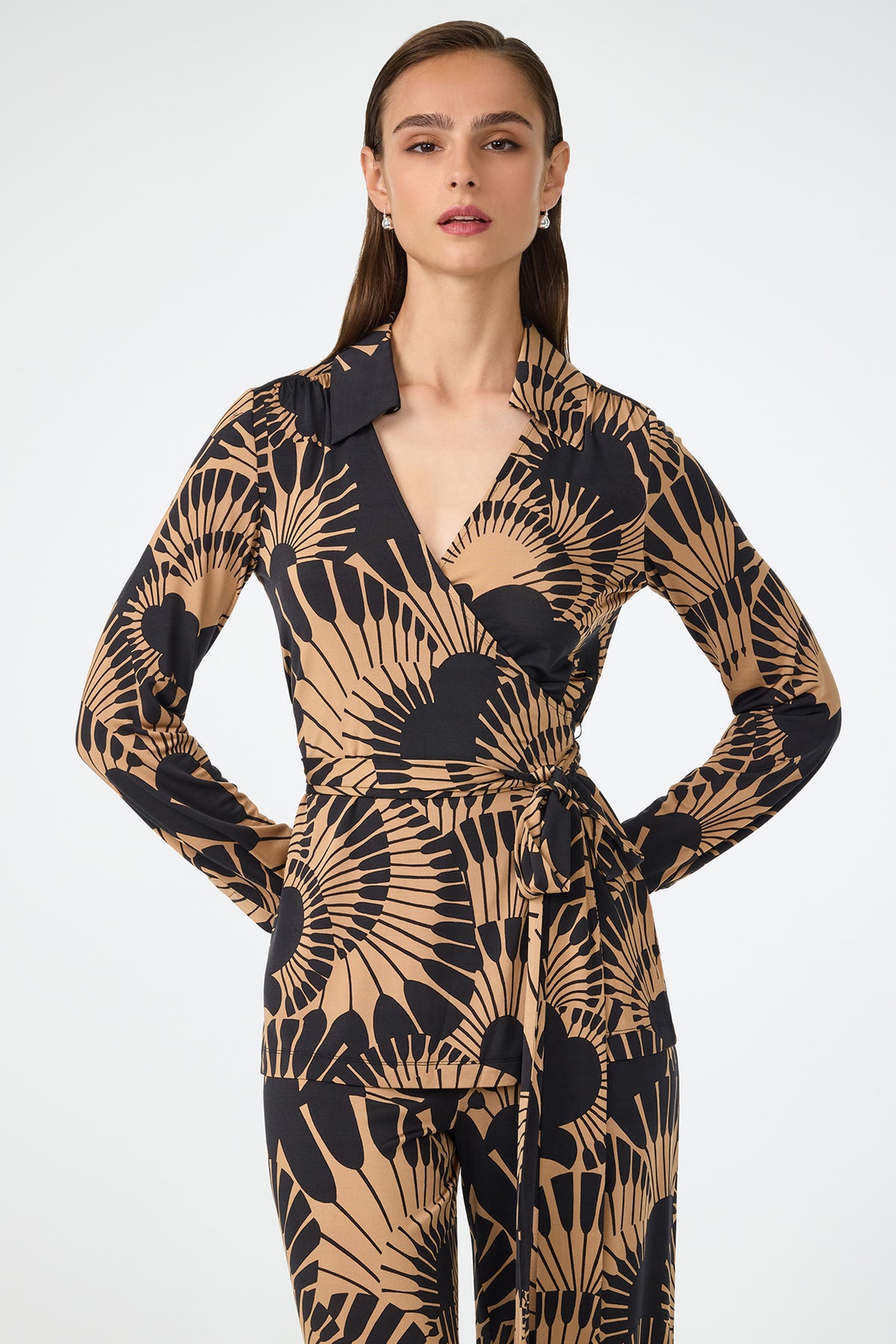 A woman with straight brown hair wears the Makeba Wrap Top in Juniper Jersey, featuring long sleeves and a matching tie at the waist, standing against a plain white background.
