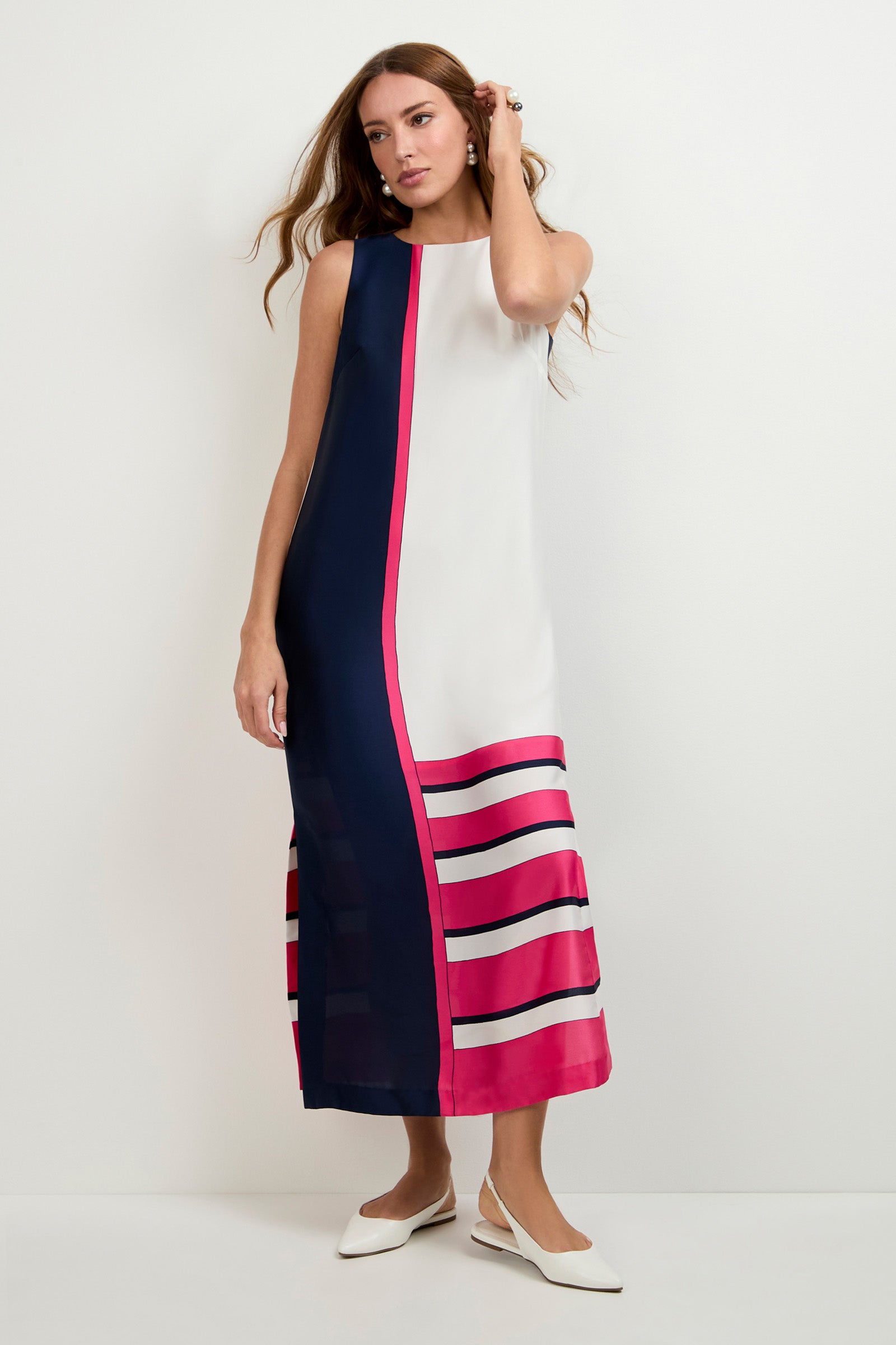 A woman stands against a plain white wall wearing the Moderna Linee Midi Dress, a sleeveless, ankle-length dress with a navy and white color block design, pink accents, and horizontal pink and navy stripes at the hem. She pairs it with white flats.