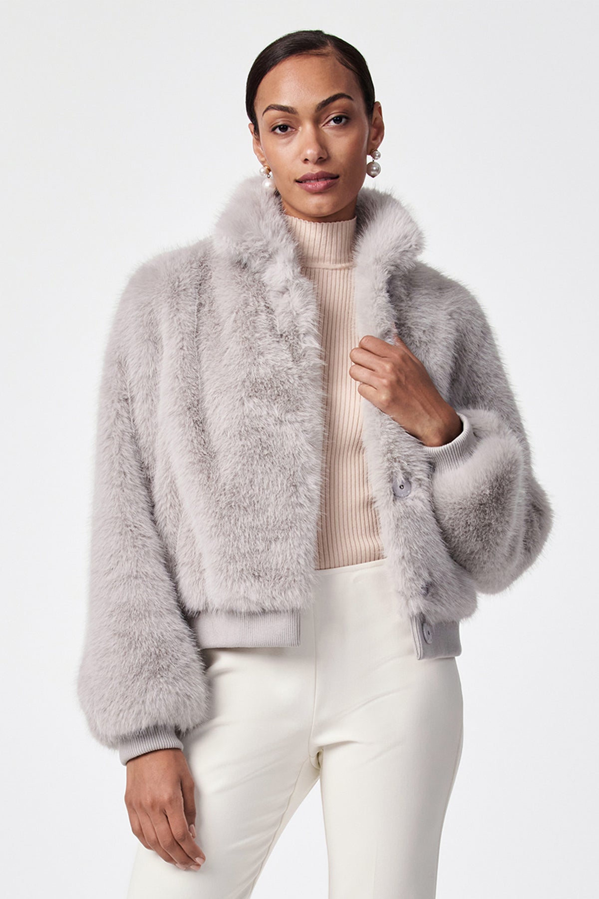 A woman stands confidently against a white background, wearing the grey Morris Short Faux Fur Coat over a beige turtleneck and white pants. Her hair is pulled back as she looks at the camera.