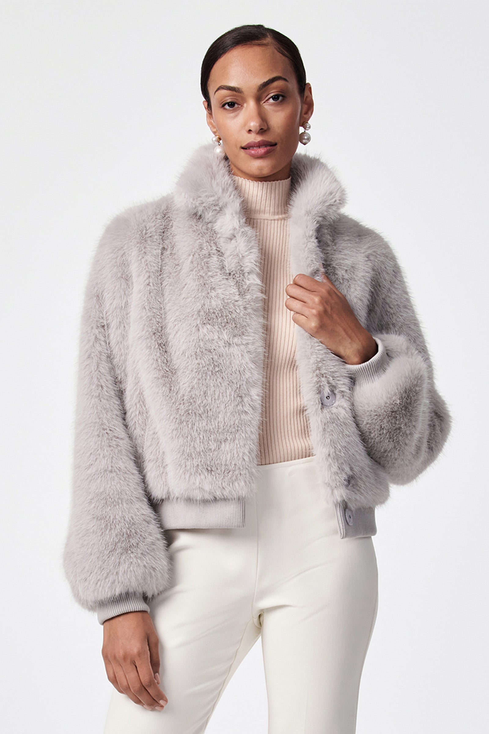 A woman stands confidently against a white background, wearing the grey Morris Short Faux Fur Coat over a beige turtleneck and white pants. Her hair is pulled back as she looks at the camera.