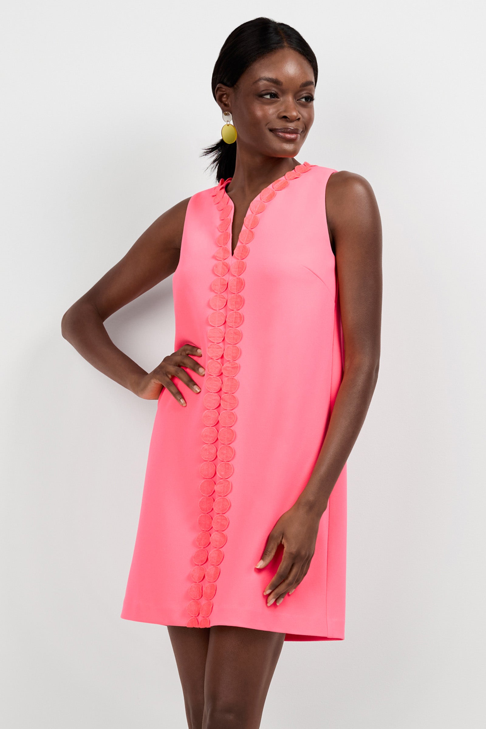 A woman stands against a plain white background, smiling with one hand on her hip, wearing the sleeveless Pavia Mini Dress in bright pink, featuring decorative circular details along the front.