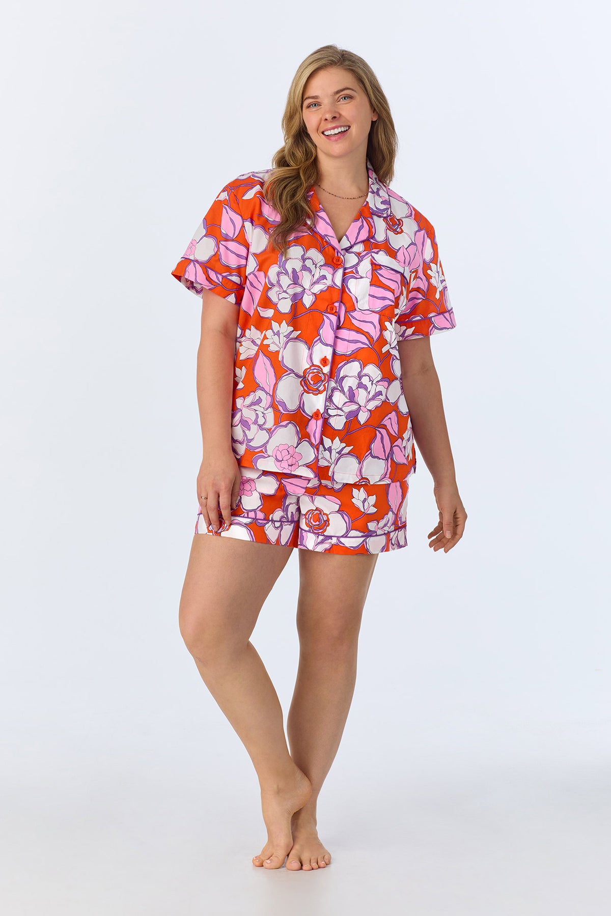 A woman with long, light brown hair stands barefoot and smiling, wearing the Shorty PJ Set - Gardenia Poplin: a colorful floral short-sleeve button-up top and matching shorts in red, pink, and white on a plain light background.