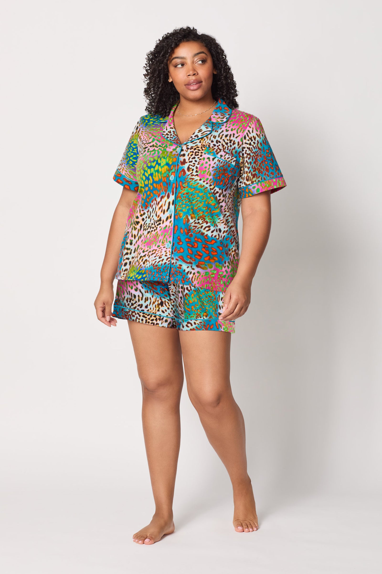 A woman stands barefoot against a white background, wearing the Shorty PJ Set - Spray Leopard: a vibrant short-sleeved button-up pajama top and matching shorts with a bold multicolored leopard print.