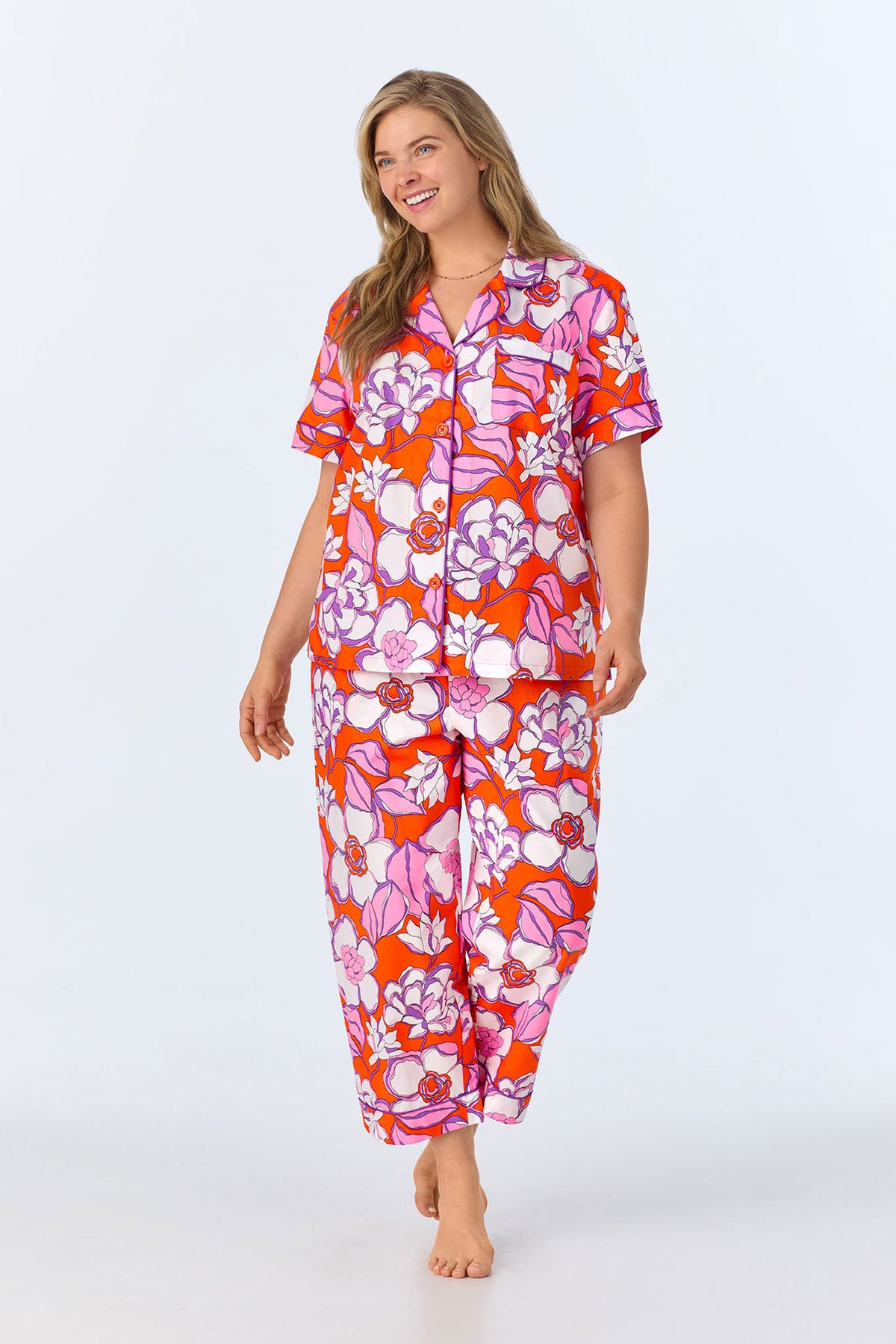 A woman smiles barefoot in the Crop Classic PJ Set - Gardenia Poplin, featuring a vibrant orange, pink, and white floral pattern on the matching short-sleeve top and pants, set against a plain light background.