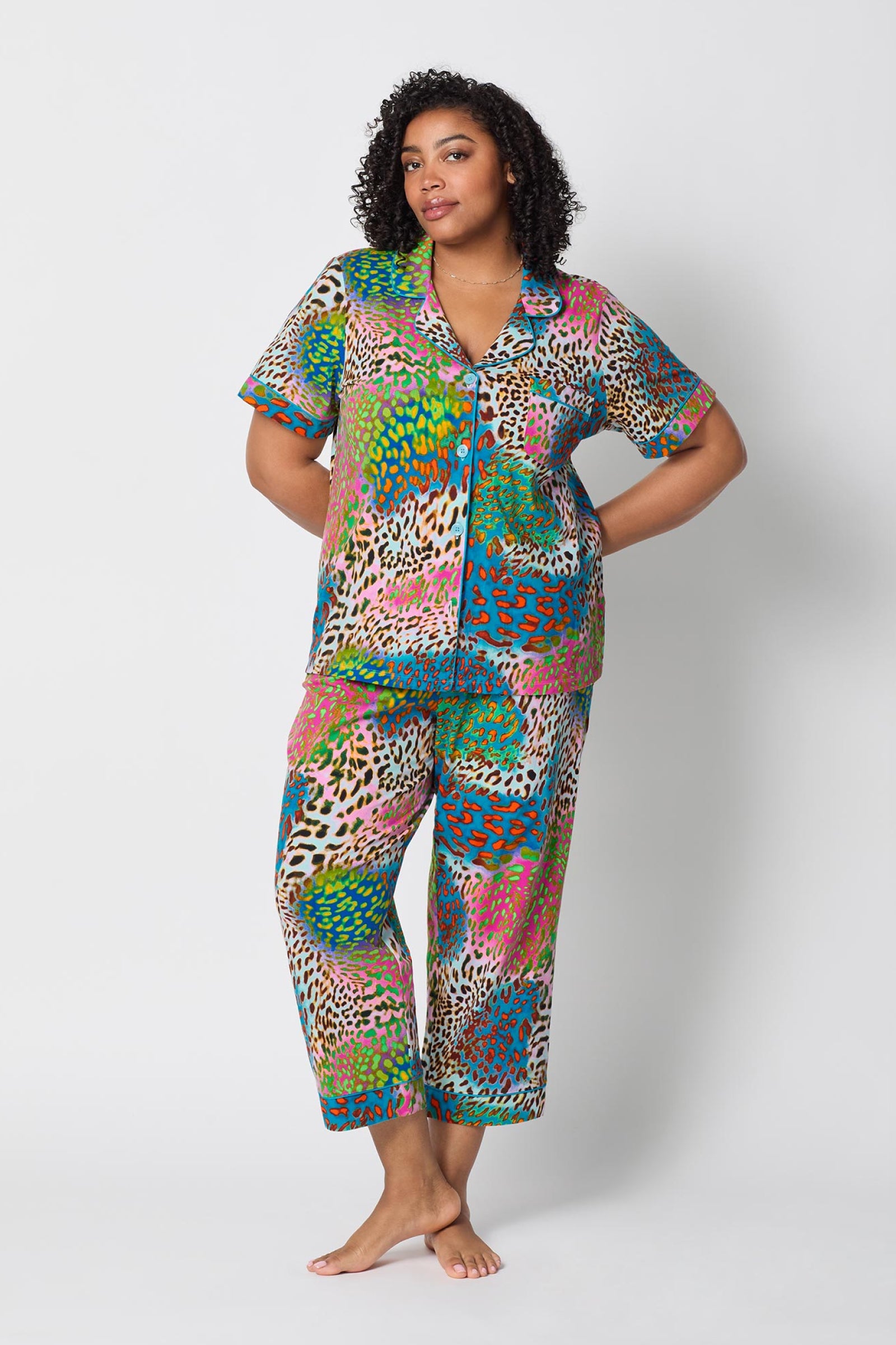 A person stands barefoot against a plain white background, wearing the Crop Classic PJ Set - Spray Leopard, which features a bold, multicolored animal print and short sleeves. They pose with one hand on their hip and a relaxed expression.