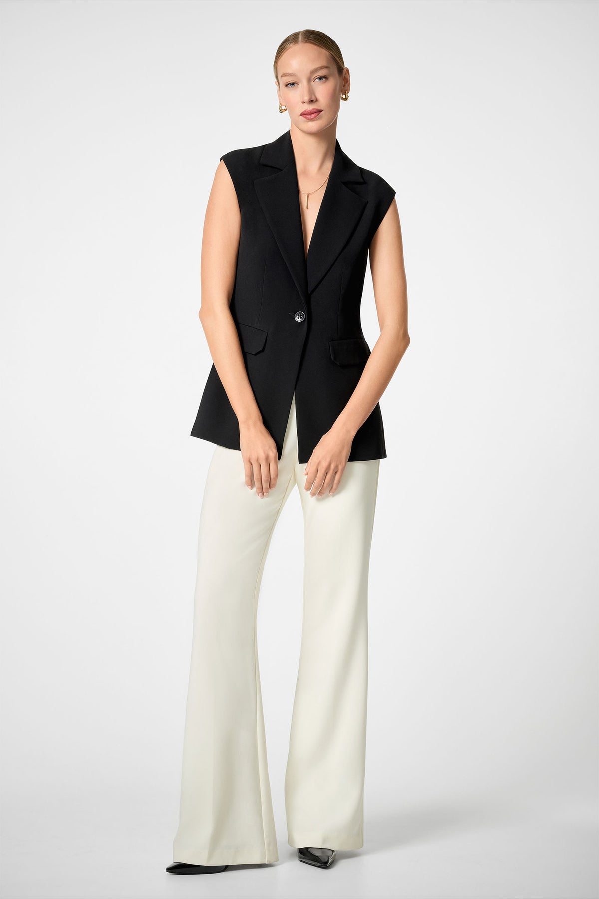 A woman stands against a plain background wearing Chimayo 2 Pants—white wide-leg trousers—paired with a sleeveless black blazer, a black top, minimal jewelry, black shoes, and her hair pulled back.