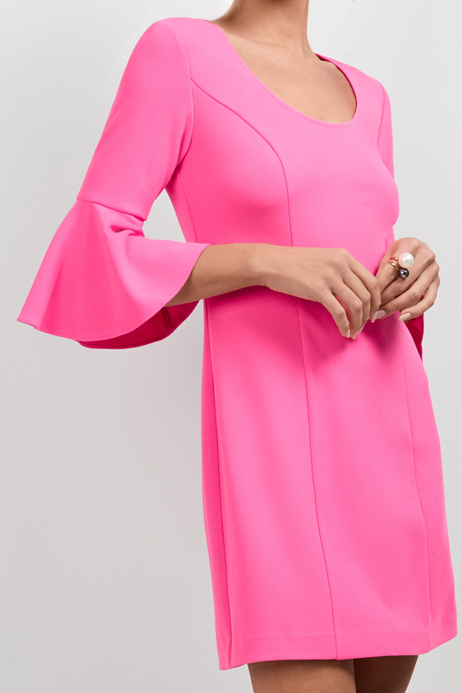 A woman wears the Scala Flounce Sleeve Dress in bright pink with long sleeves and flared cuffs, standing against a plain light background with her hands clasped. Her face is partially out of frame.