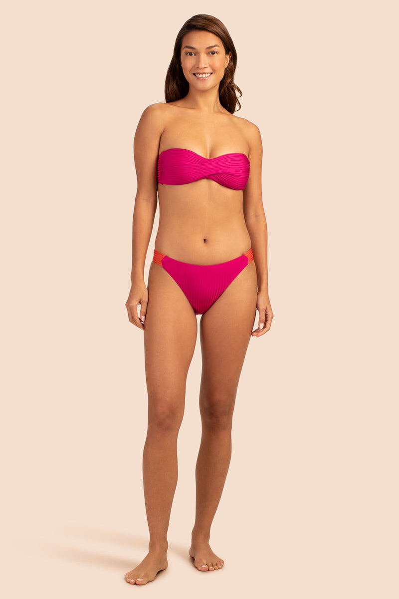 A woman stands smiling in a strapless pink bandeau bikini top and Women's Olympia Rib Low Rise French Cut Swim Bottom, both with textured fabric, against a plain beige background.